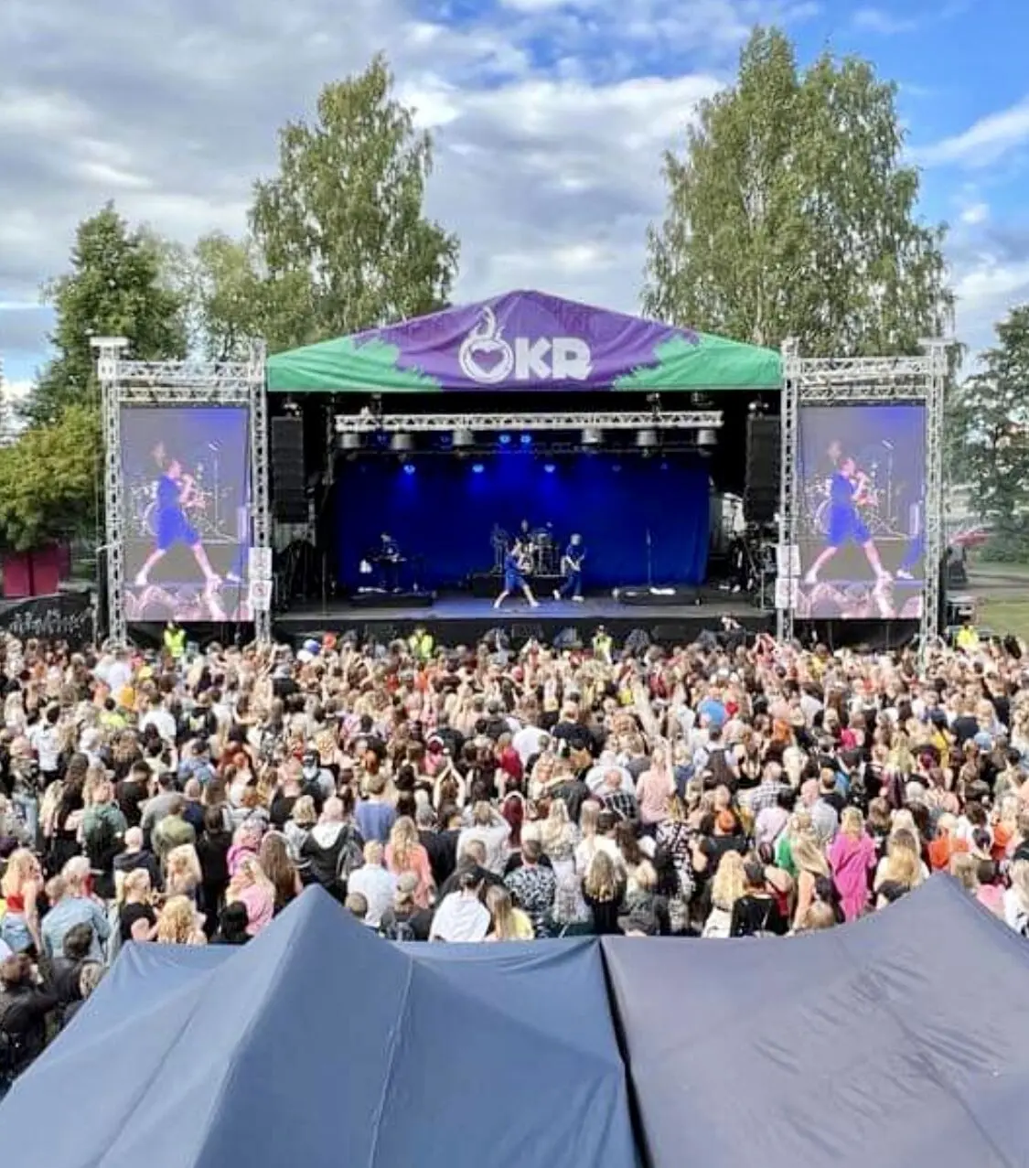 Katettu festivaalilava, LED-näytöt ja yleisö puiden keskellä